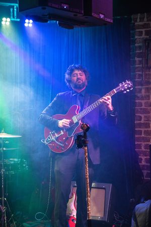 A musician performs on stage with a red guitar under dramatic blue and purple stage lighting.