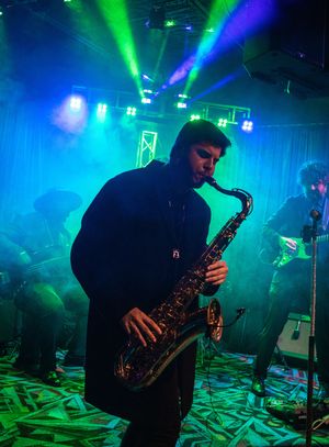 Saxophone player performs on stage in dark venue with dramatic blue and green lighting creating atmospheric ambiance.