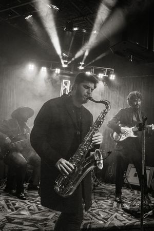 Musicians perform with saxophones and instruments in a moody, atmospheric venue with dramatic lighting and smoke effects.