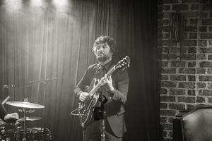 A musician performs on stage with a guitar under spotlights against a brick wall backdrop in black and white.