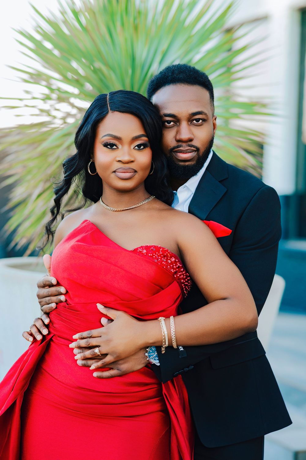 Couple poses for romantic outdoor photos with palm trees, wearing red gown and black suit.
