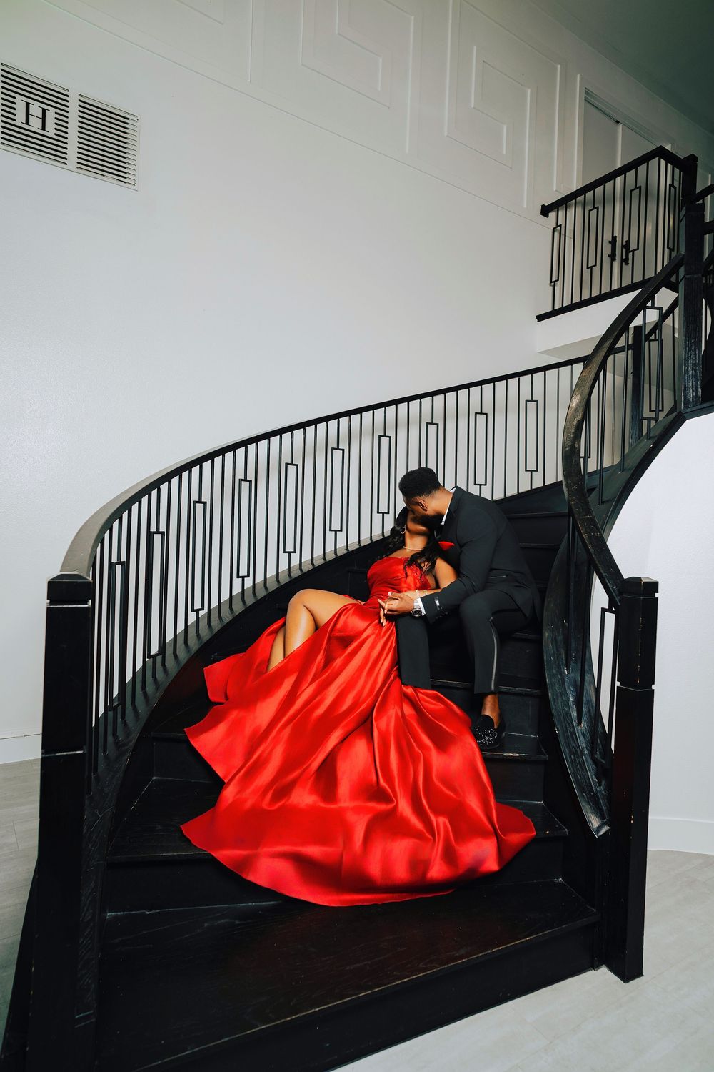 A couple in formal attire pose dramatically on an elegant black spiral staircase, with the woman wearing a flowing red gown.