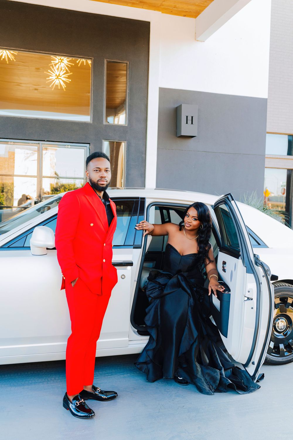 A stylish couple in formal wear pose next to a white luxury car, with one person wearing a bold red suit.