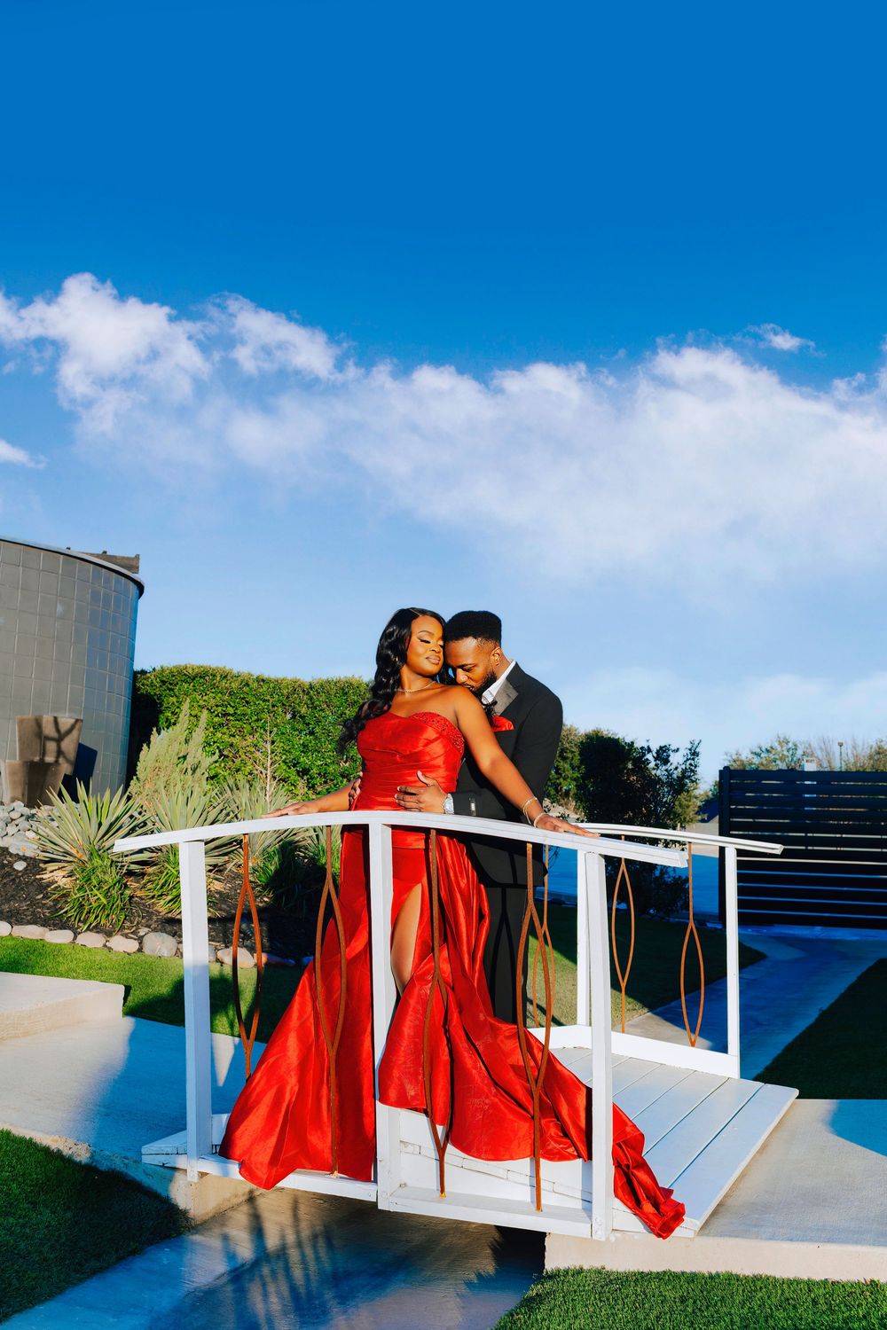 A couple in a red dress poses romantically against white railings on a rooftop terrace with blue sky views.