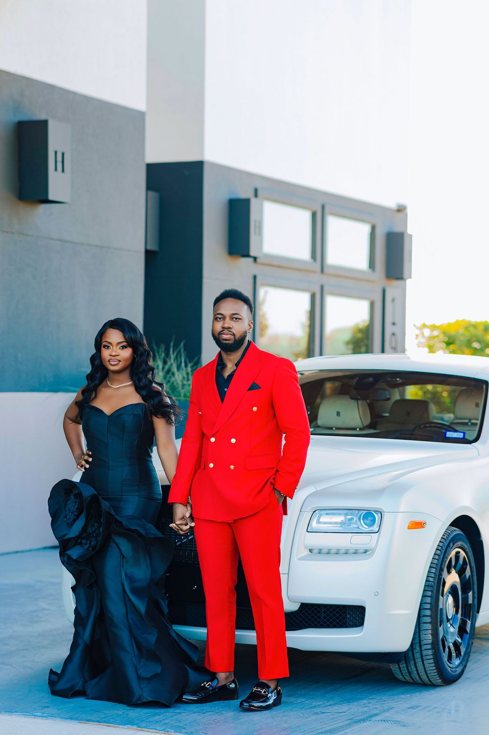 A couple in elegant evening wear stand beside a white Rolls Royce, featuring a striking red suit and navy gown.