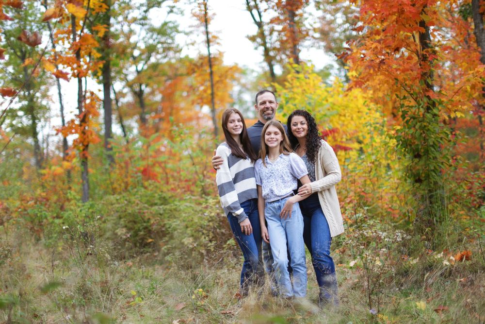 A group of friends laugh and embrace while posing for autumn family photos among vibrant orange fall foliage.