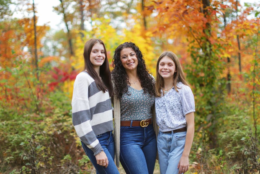 Three people pose together outdoors with autumn foliage and trees creating a colorful fall backdrop.