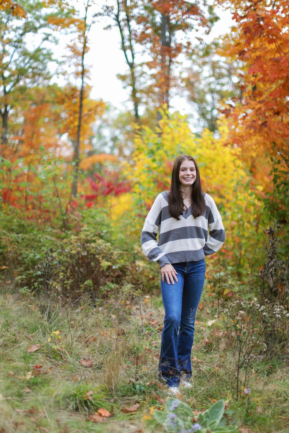Person in striped sweater and jeans poses outdoors against colorful fall foliage backdrop.