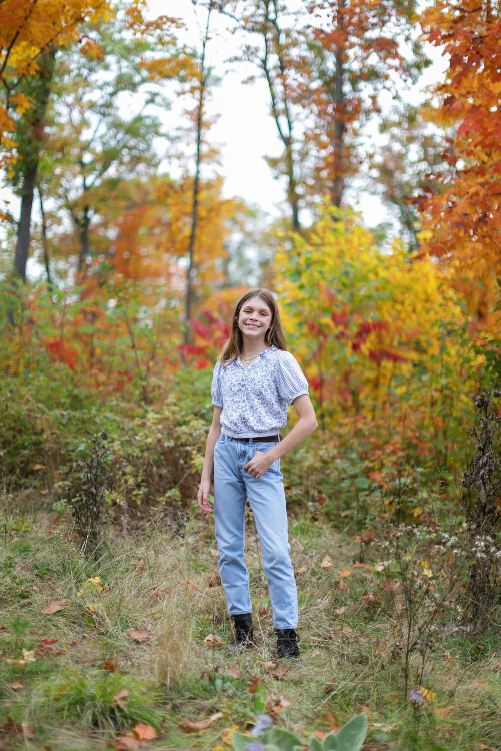 Person in casual attire poses in a meadow surrounded by autumn trees with orange and yellow leaves.
