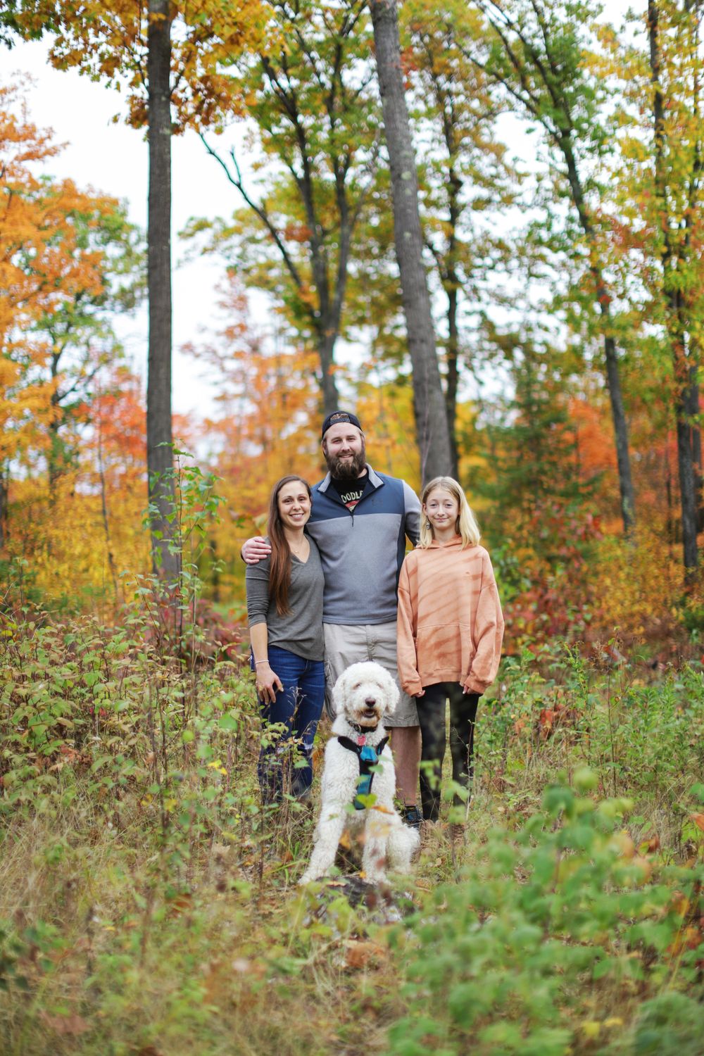 A group walks together with their fluffy white dog through a scenic autumn forest with golden foliage.