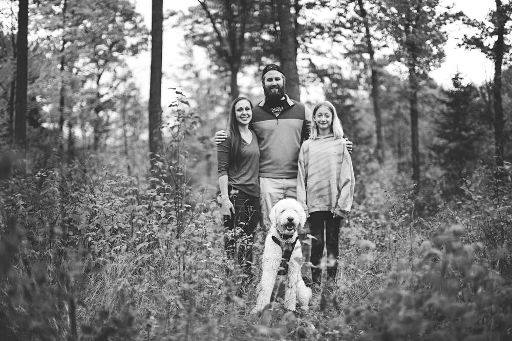 A group of people and their dog pose together on a wooded path during an autumn day.
