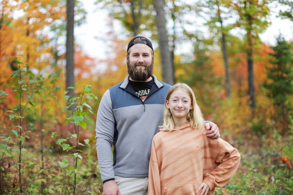 A couple stands together smiling outdoors surrounded by vibrant autumn foliage in a forest setting.