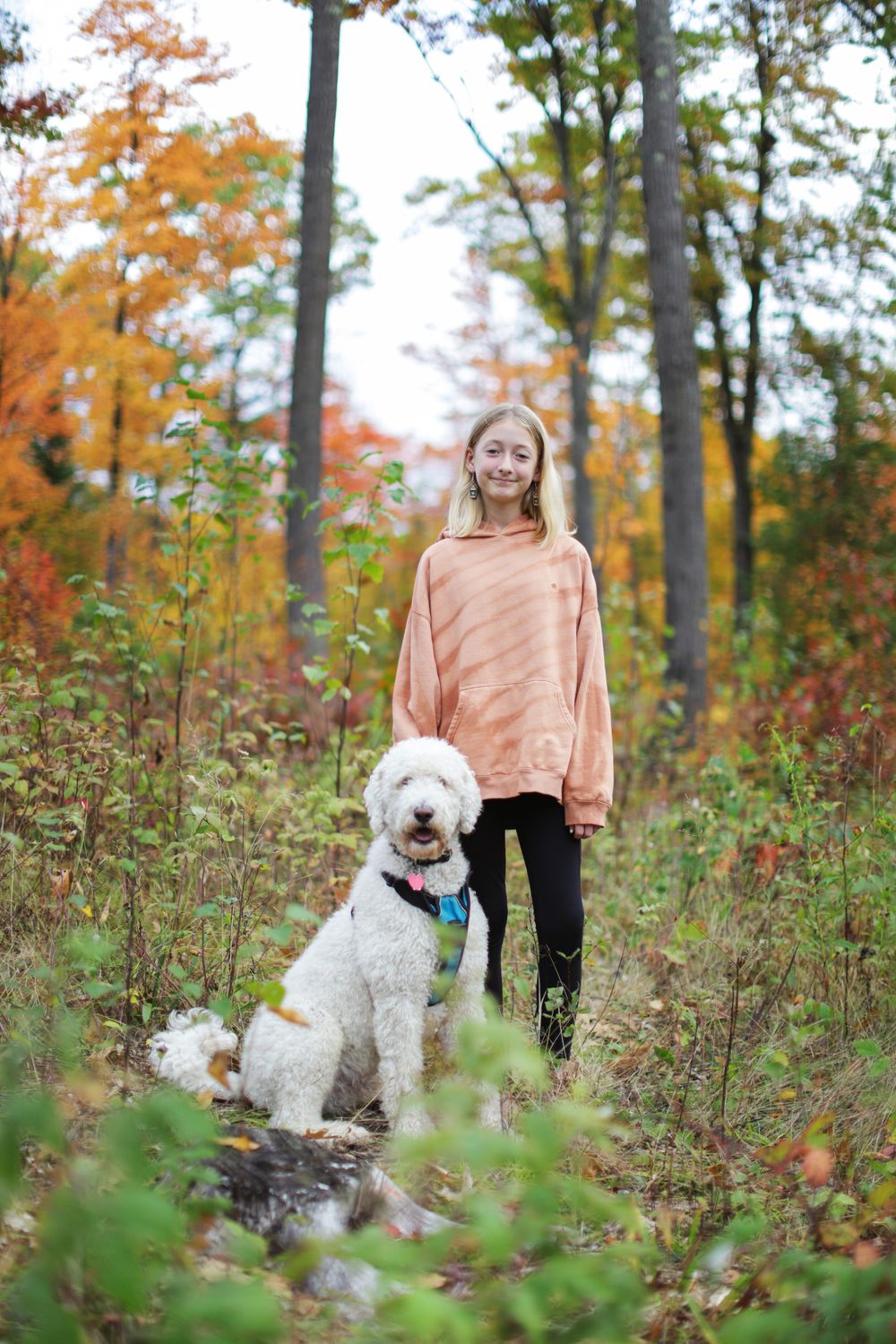 Two people pose with their fluffy white dogs among fall foliage in a wooded setting.