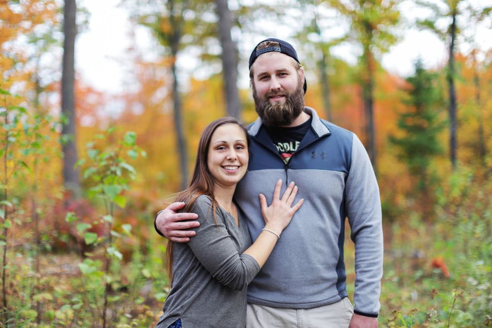 A couple poses together outdoors during autumn with colorful fall foliage in the background.
