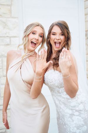 Two women in wedding attire share joyful laughter while posing together in front of a white stone wall.