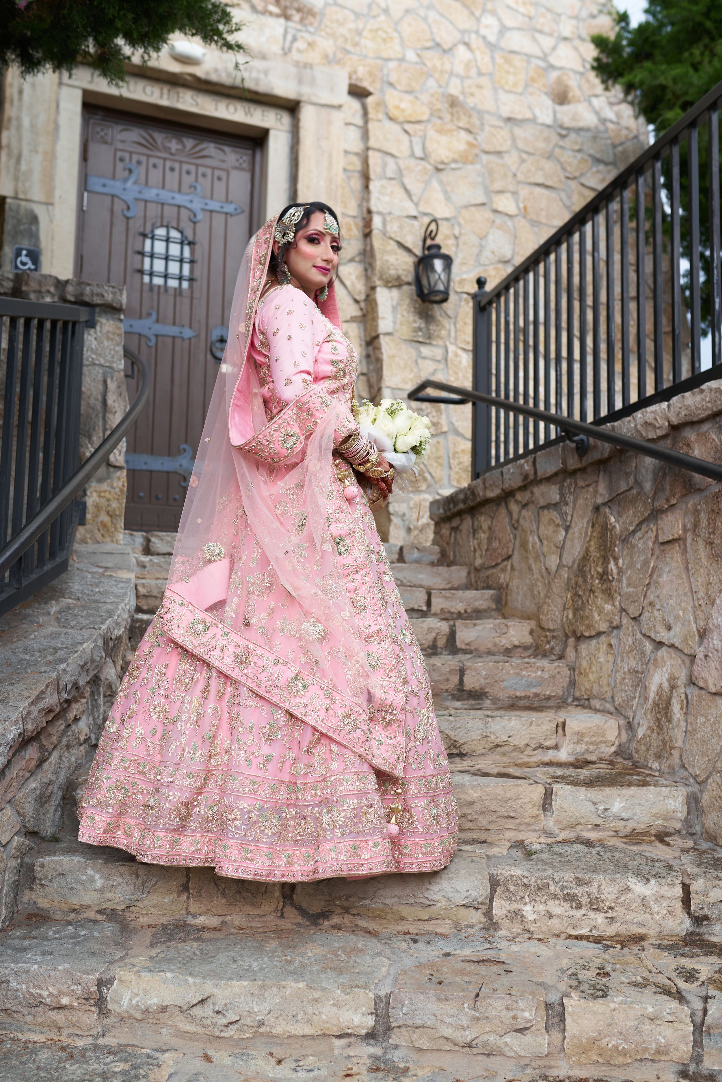 A bride in an ornate pink lehenga with floral embroidery stands on stone steps near a wooden door.