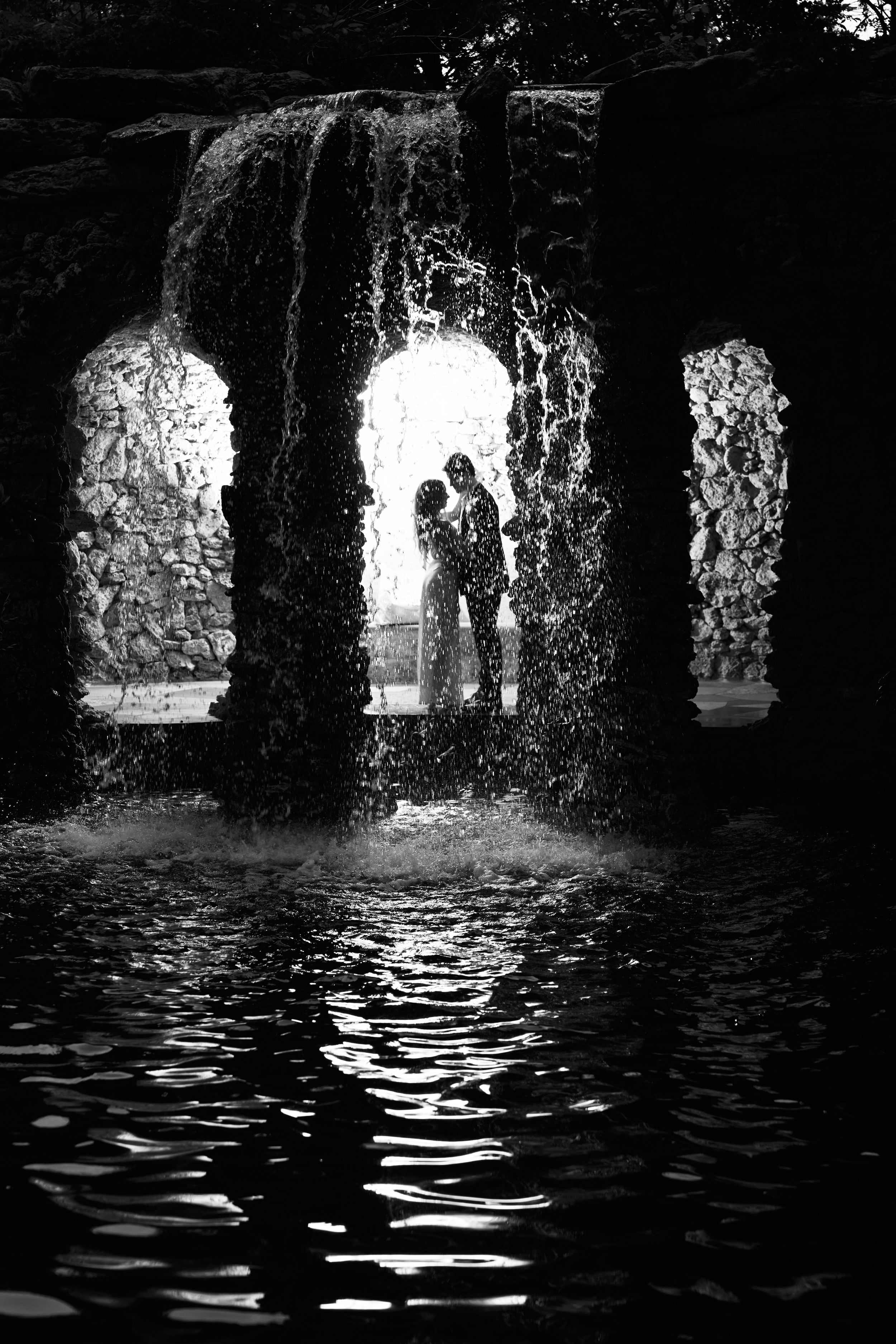 Black and white photo of stone archways reflecting on rippling water below.