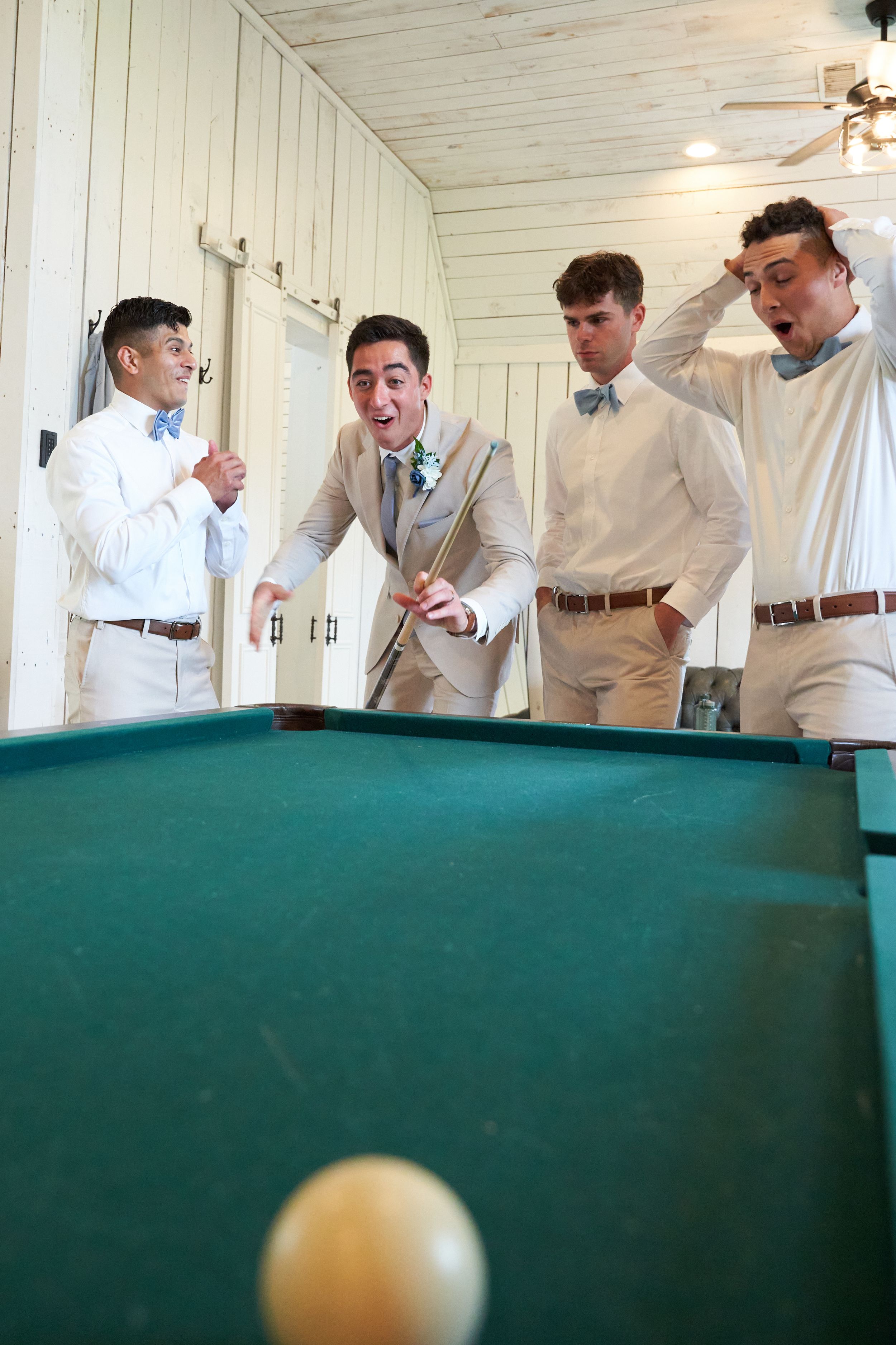 Groomsmen in light colored suits gather around a pool table during wedding preparations.