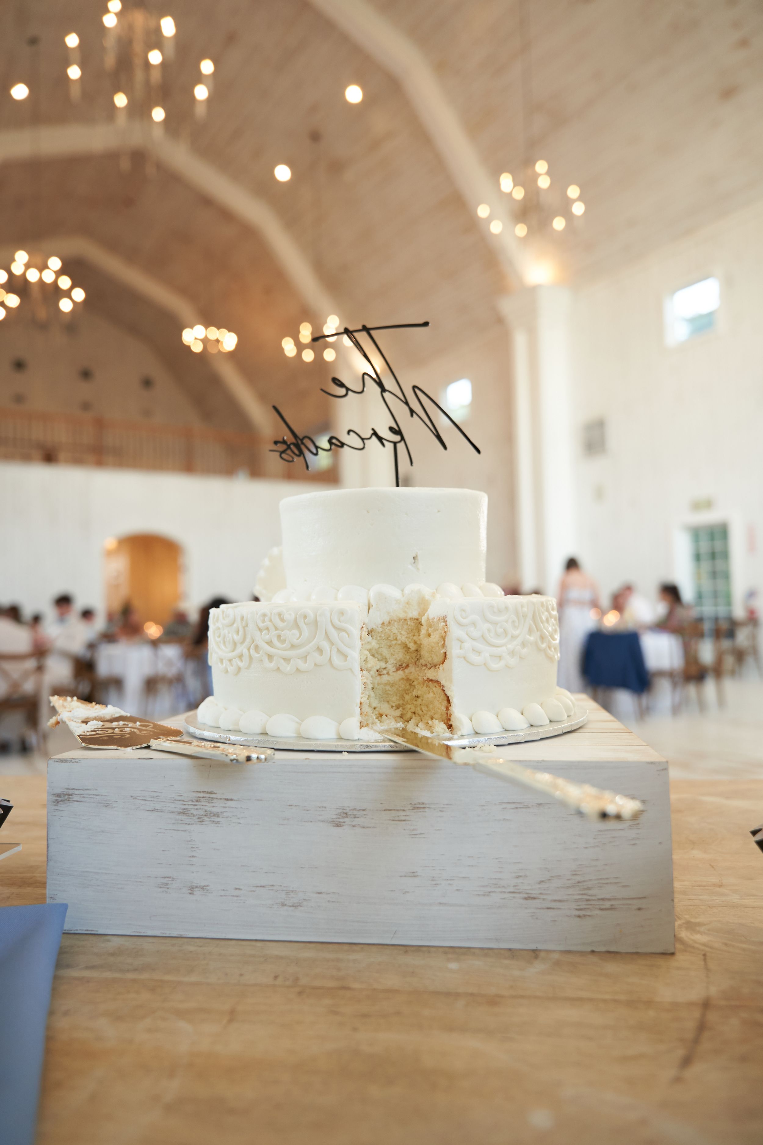 A white wedding cake with delicate lace details sits on a rustic wooden stand in an elegant venue with twinkling lights.