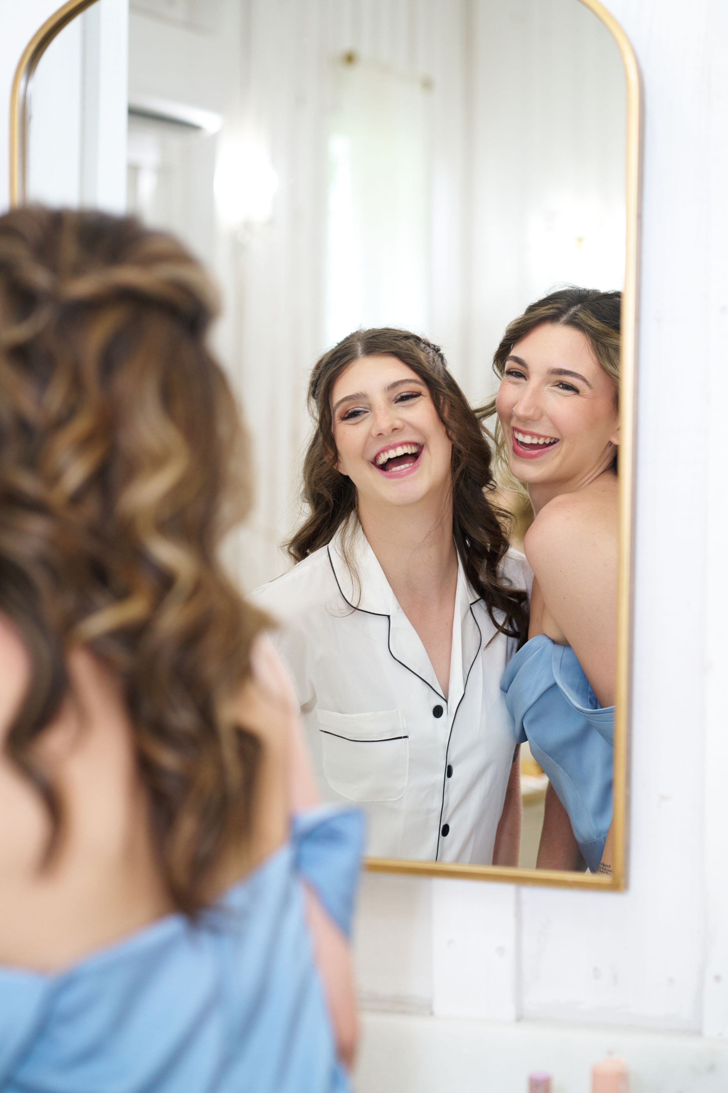 Two women in robes share laughter while looking at their reflection in a gold-framed mirror.