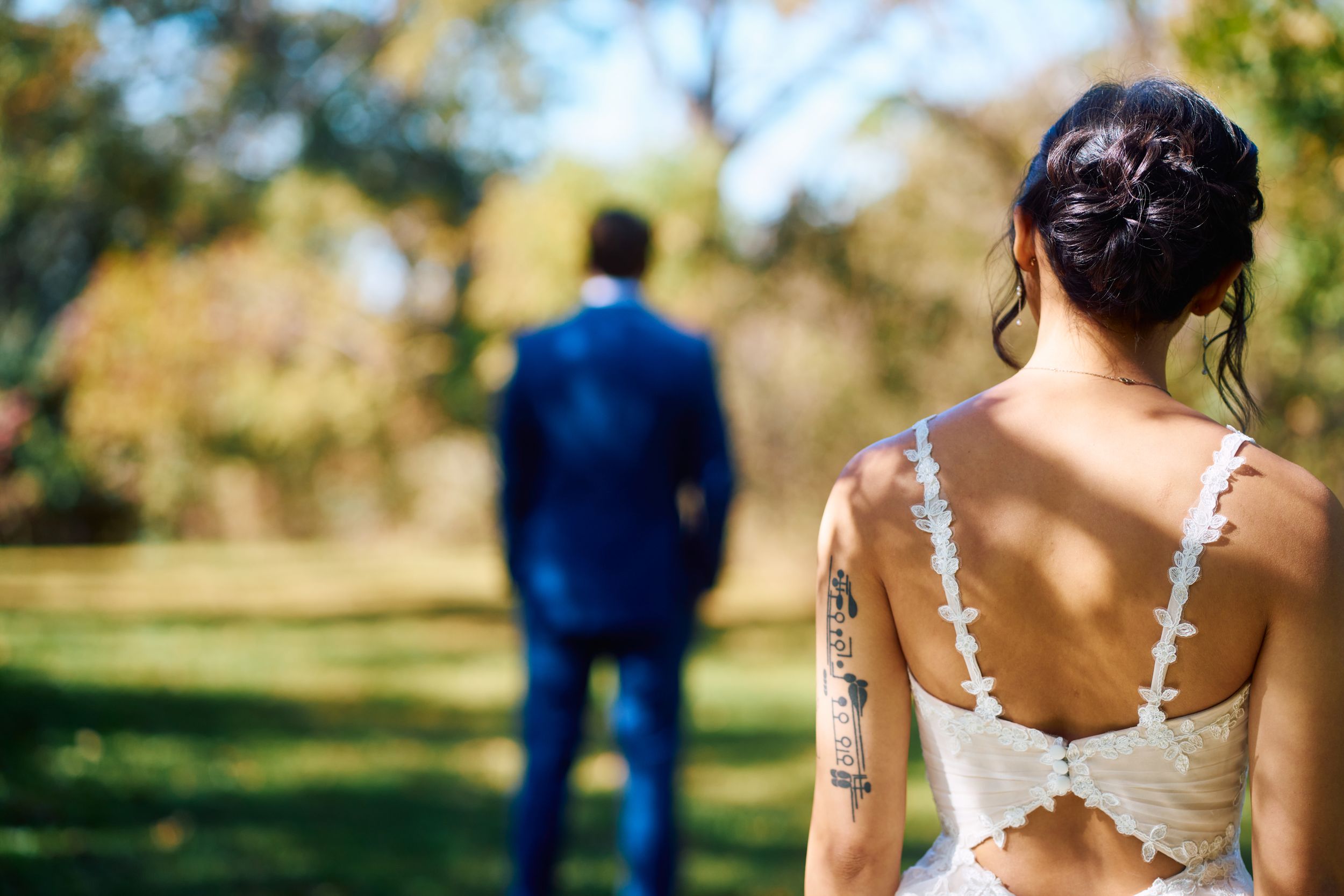 A bride in a lace back dress faces away while her groom stands in the distance outdoors.