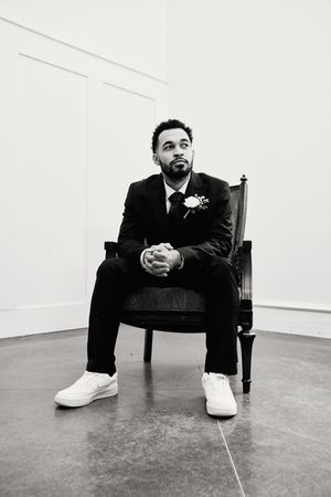 A person in a formal suit and white sneakers sits contemplatively on a wooden chair in a minimalist setting.