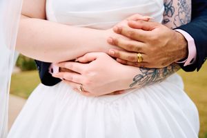 Close-up details of hands and wedding rings against white dress fabric showing delicate tattoo artwork.
