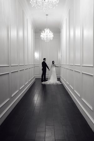 A black and white photo of newlyweds standing in an elegant hallway with crystal chandeliers.