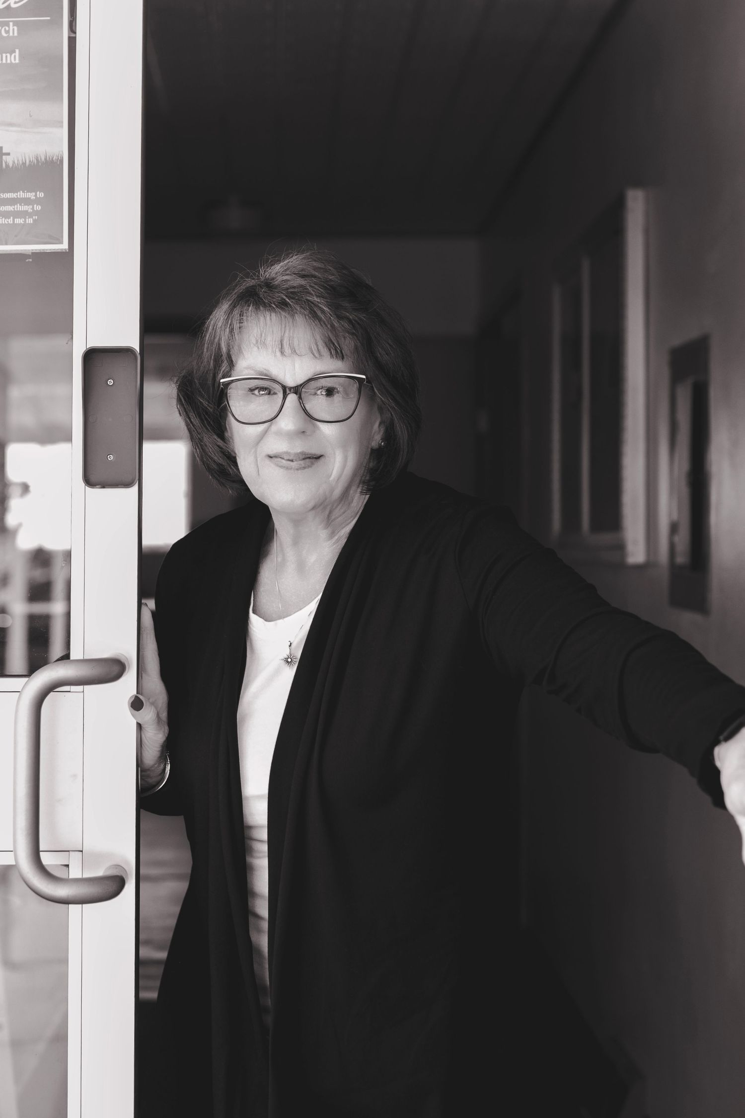 Black and white photo of woman in glasses holding open a glass door.