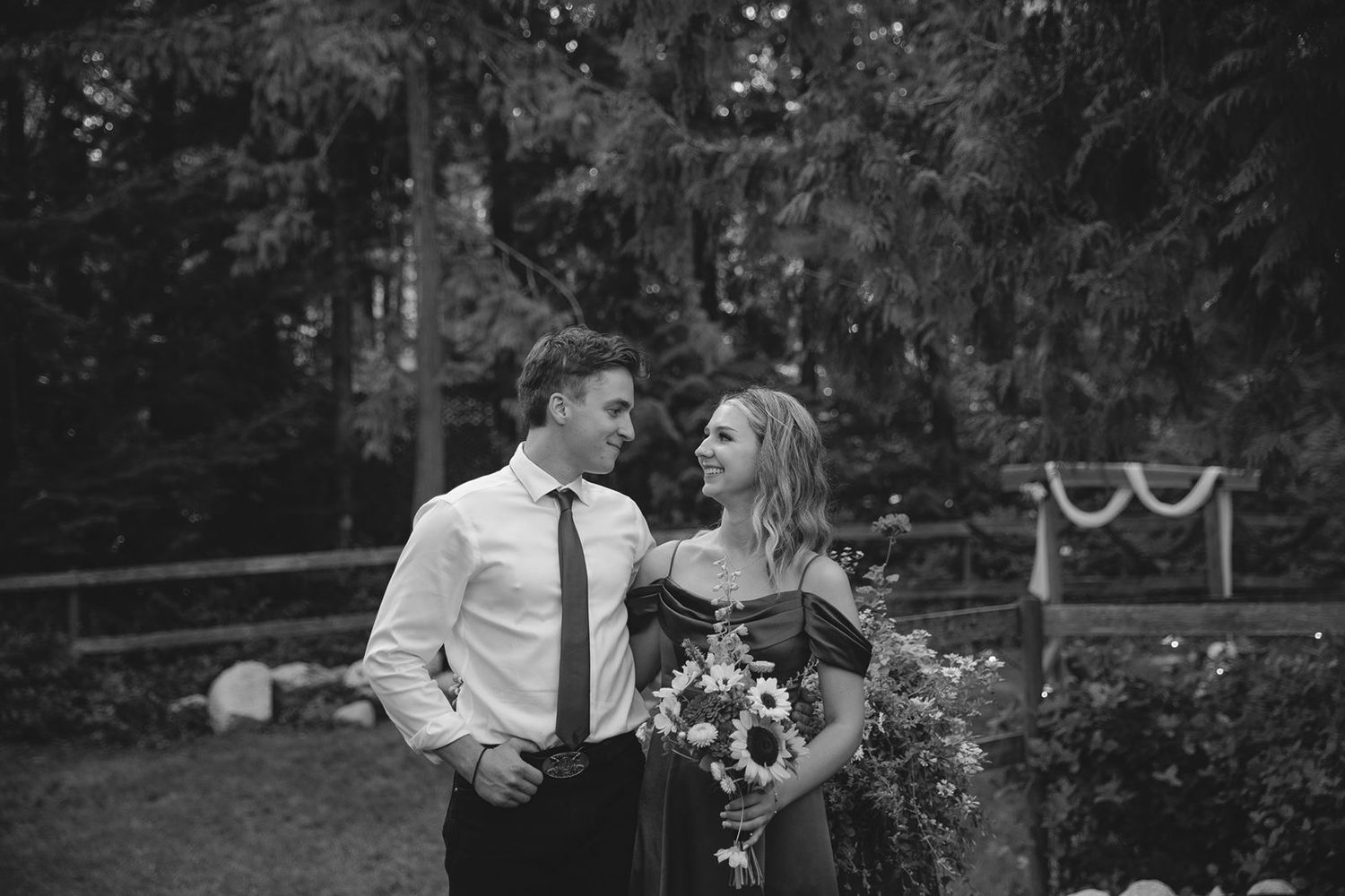 Black and white photo of a couple sharing a romantic moment outdoors at a wedding venue surrounded by trees.