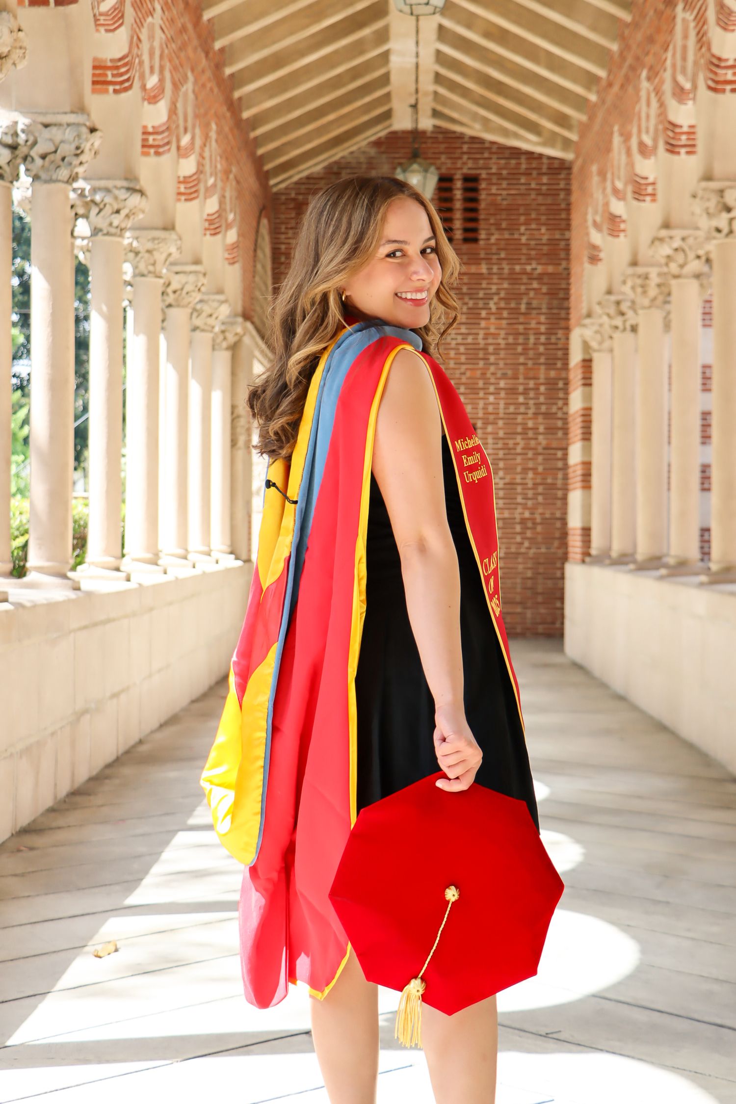 A person in a black swimsuit and colorful red cape walks through an elegant columned hallway with sunlit arches.
