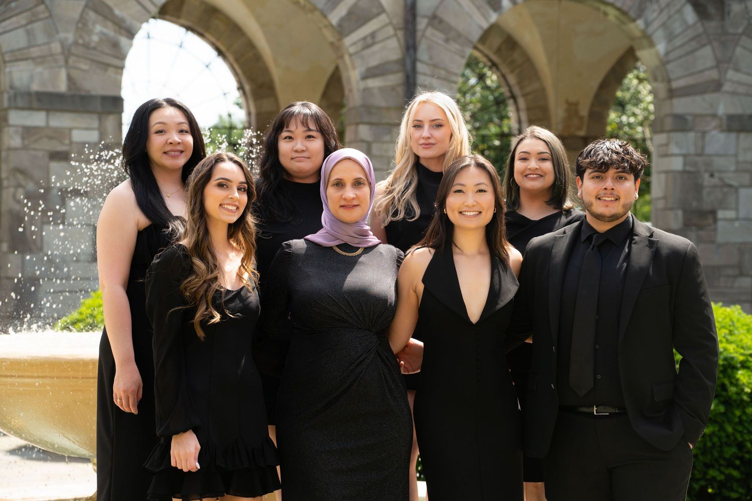 Group of people wearing formal black attire pose together under a stone archway in an outdoor setting.