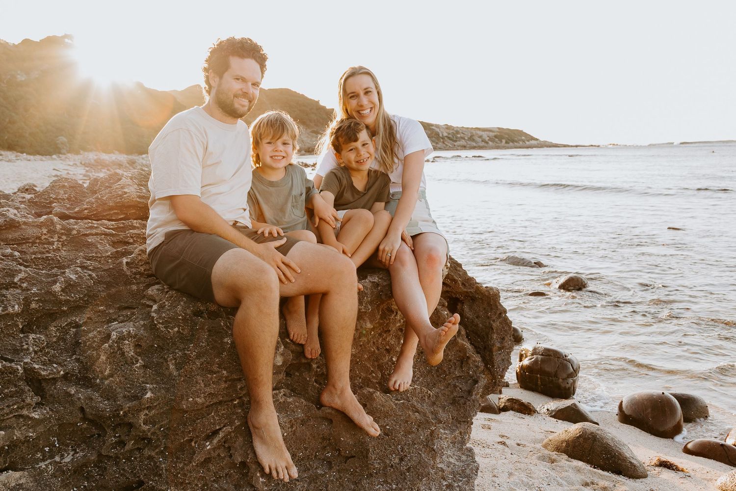 Family and dog enjoying a sunset on rocky beach shore while waves crash in the background.