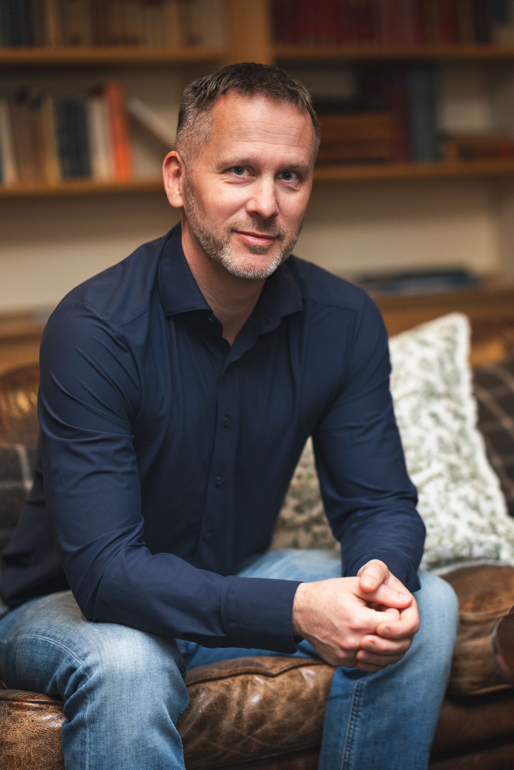 A man in a navy blue shirt sits casually on a brown leather couch in a room with bookshelves in the background.
