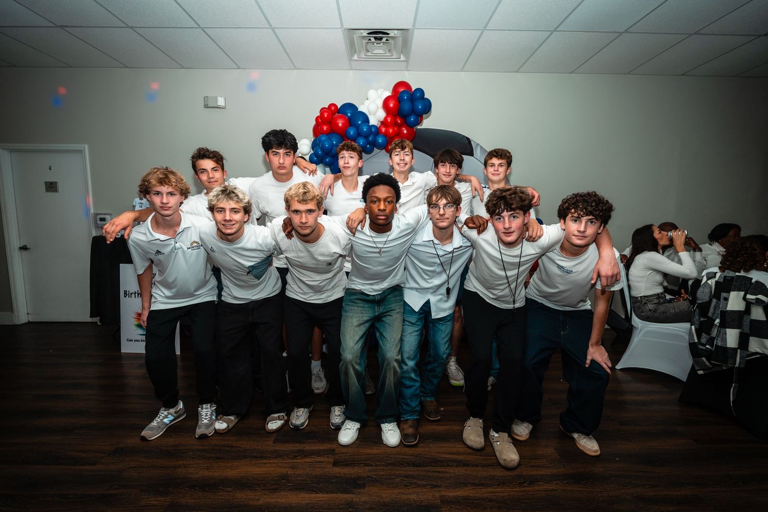 A group in white shirts and jeans pose together against a wall in a casual indoor setting.