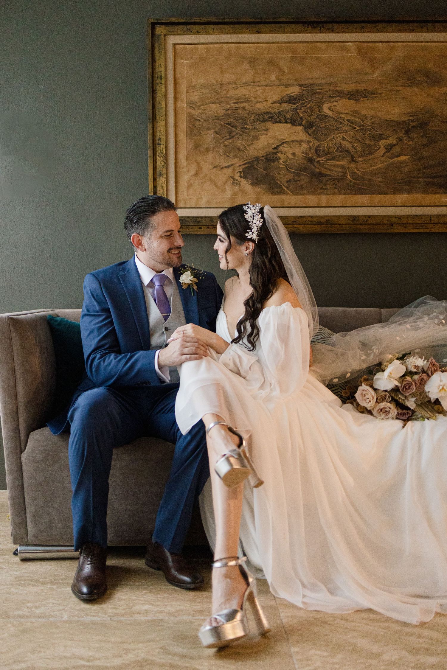 Una romántica pareja de novios con traje azul y vestido blanco se sienta junta en un elegante sofá en una habitación vintage.