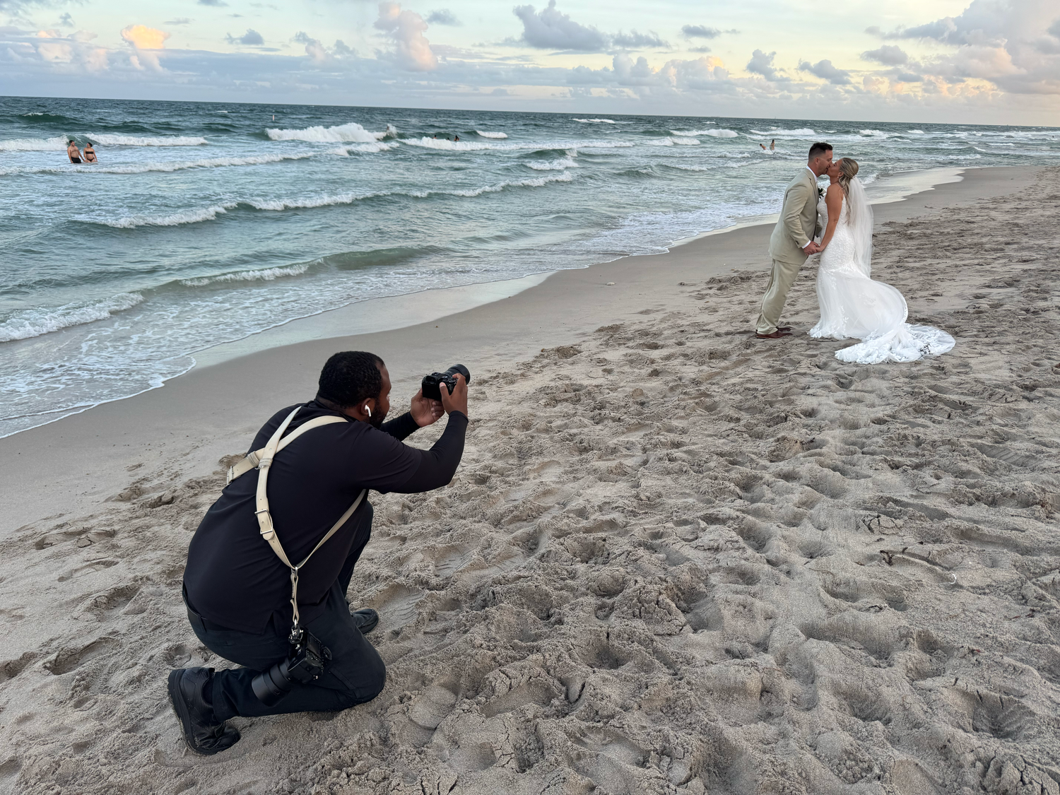 Professional photographer captures a couple during a romantic beach wedding photoshoot at sunset with waves crashing behind them.