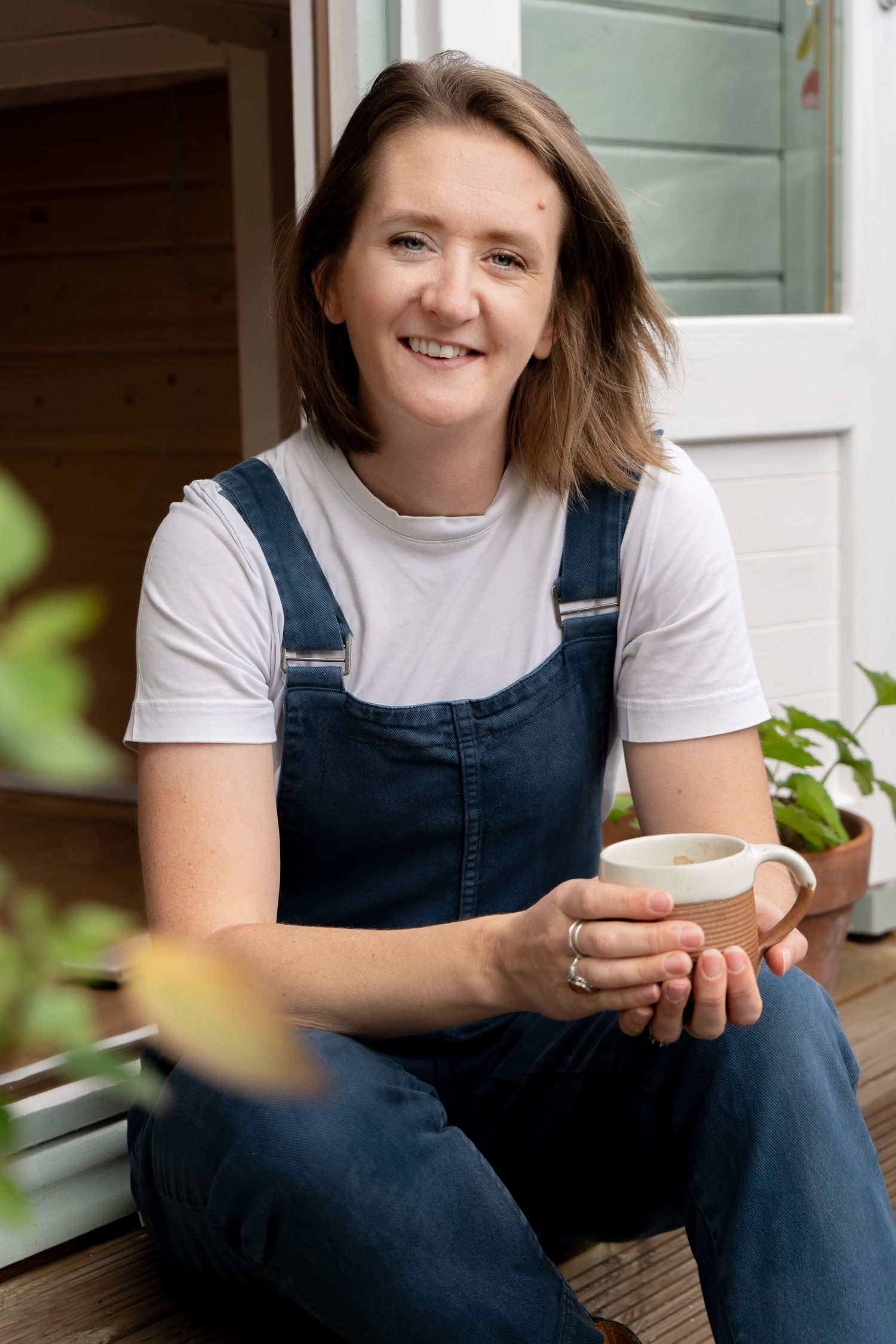 Person in denim overalls and white t-shirt sits relaxed with coffee cup in hand.