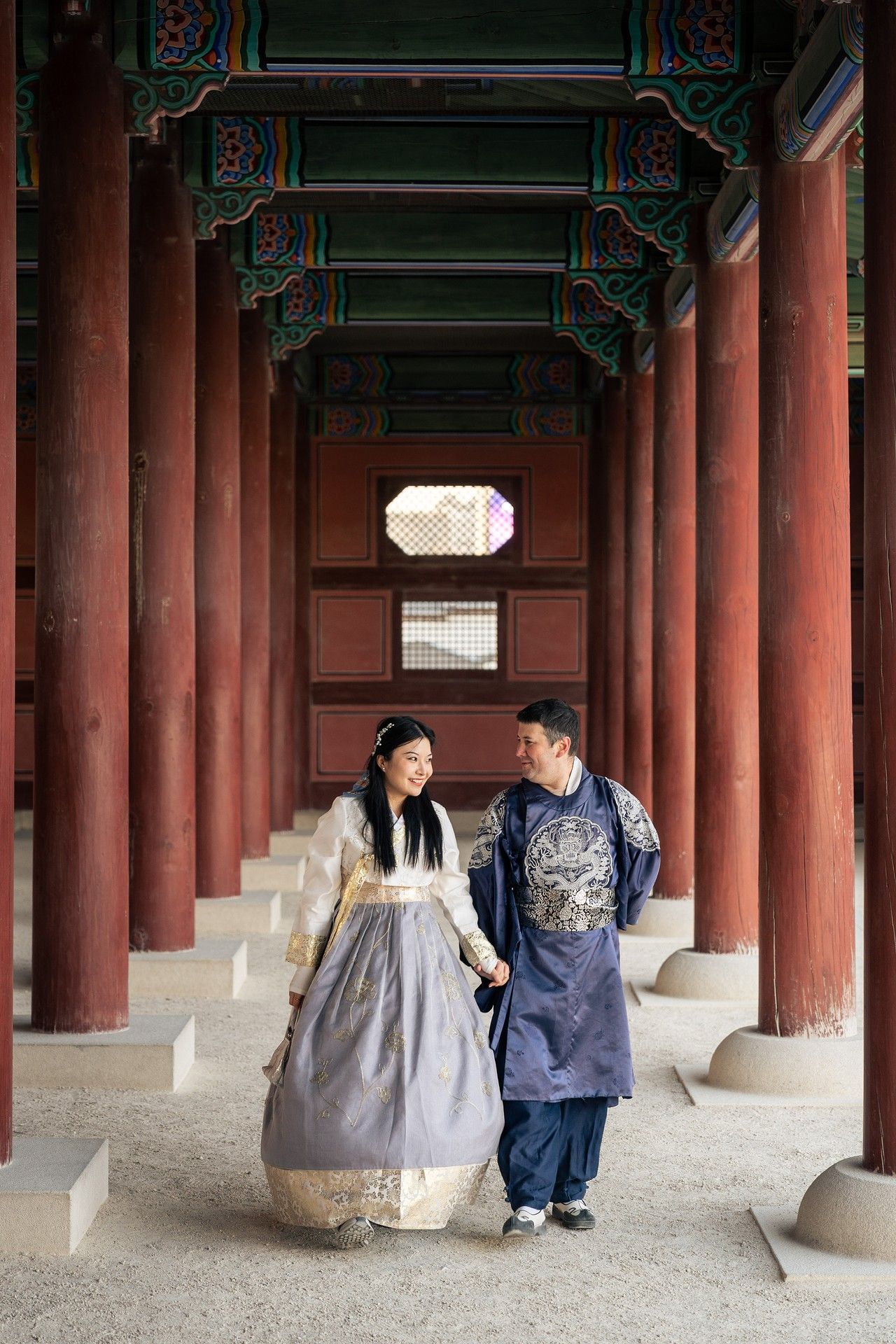 A couple in traditional Korean hanbok attire walking through a historic palace corridor with red pillars and ornate ceiling.