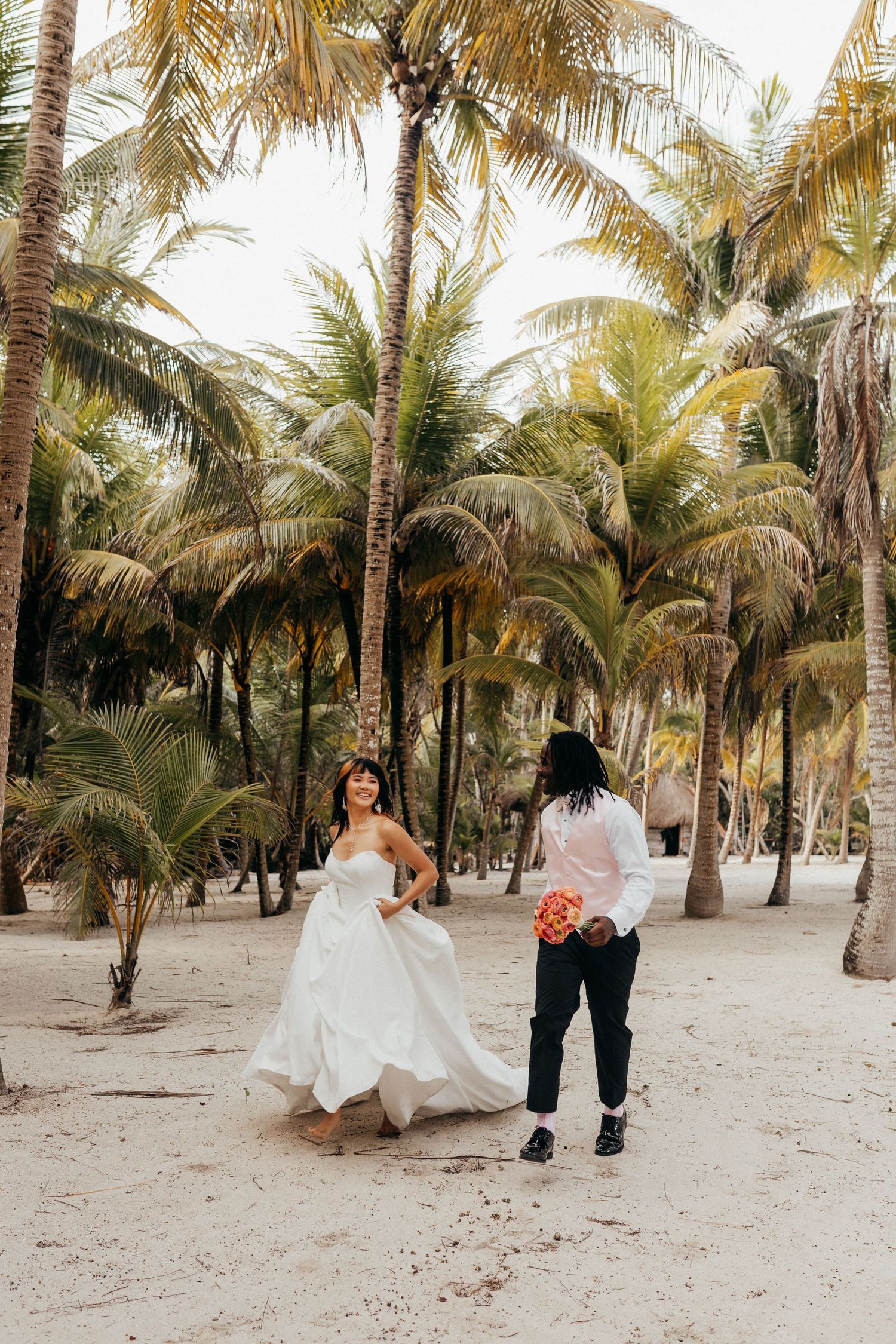 A couple walks through a scenic palm tree grove on a sandy beach during a tropical wedding photoshoot.