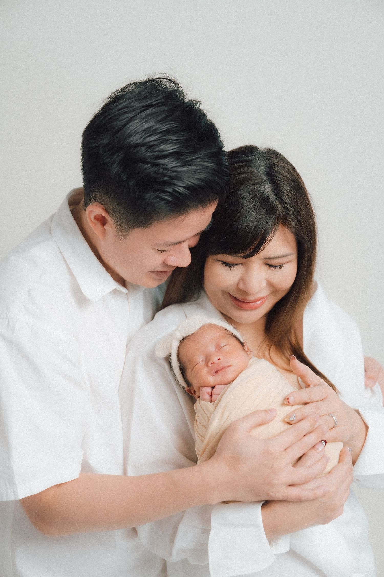 Family in white clothing sharing tender moment with newborn on light background.