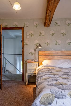 Cozy bedroom with floral wallpaper, exposed wooden beam, and rustic headboard with bedside table.