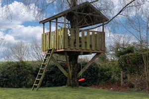 Wooden treehouse platform with ladder in garden under blue sky.