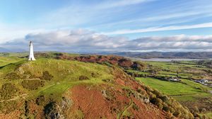 Panoramic coastal landscape with rolling green hills, cliffs and lighthouse overlooking the ocean under blue skies.