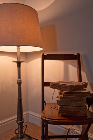 Vintage table lamp illuminating stack of old books on wooden chair in dimly lit room.