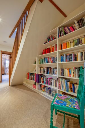 Built-in bookshelf under stairs with colorfully organized books and a turquoise accent chair.