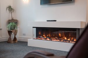 Modern linear electric fireplace with realistic flames mounted in a sleek white surround with indoor plants nearby.