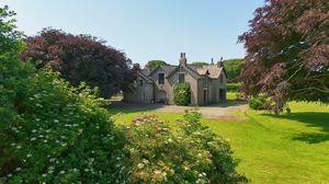 Historic stone house with lush gardens and mature trees under bright blue summer sky.