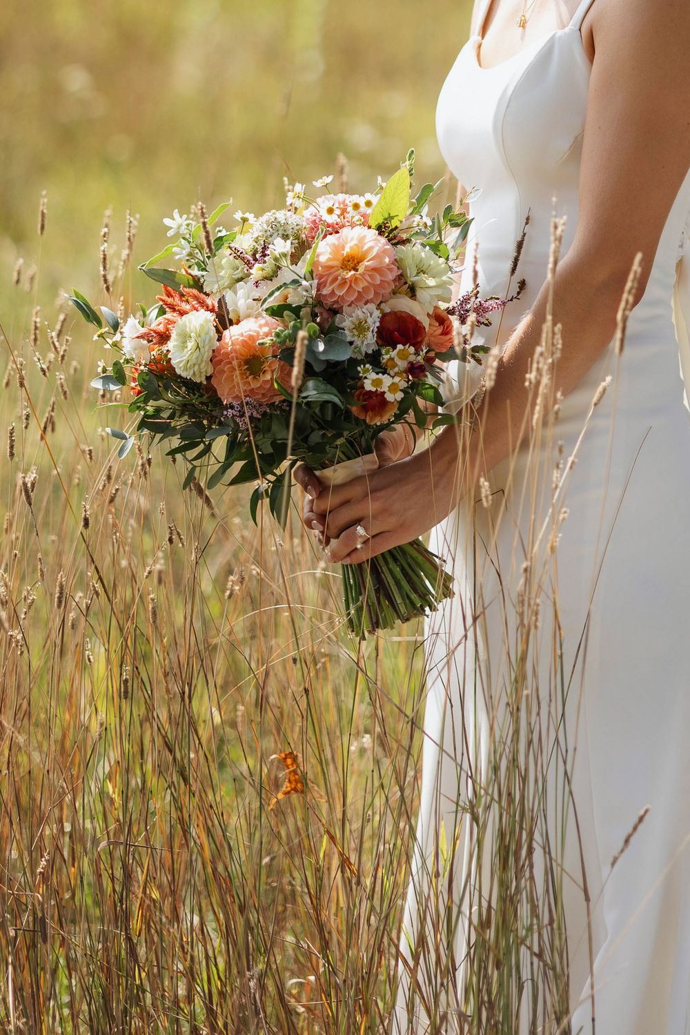 A bride holds a beautiful peach and coral floral bouquet against tall grass in a natural setting upsy-daisy floral.