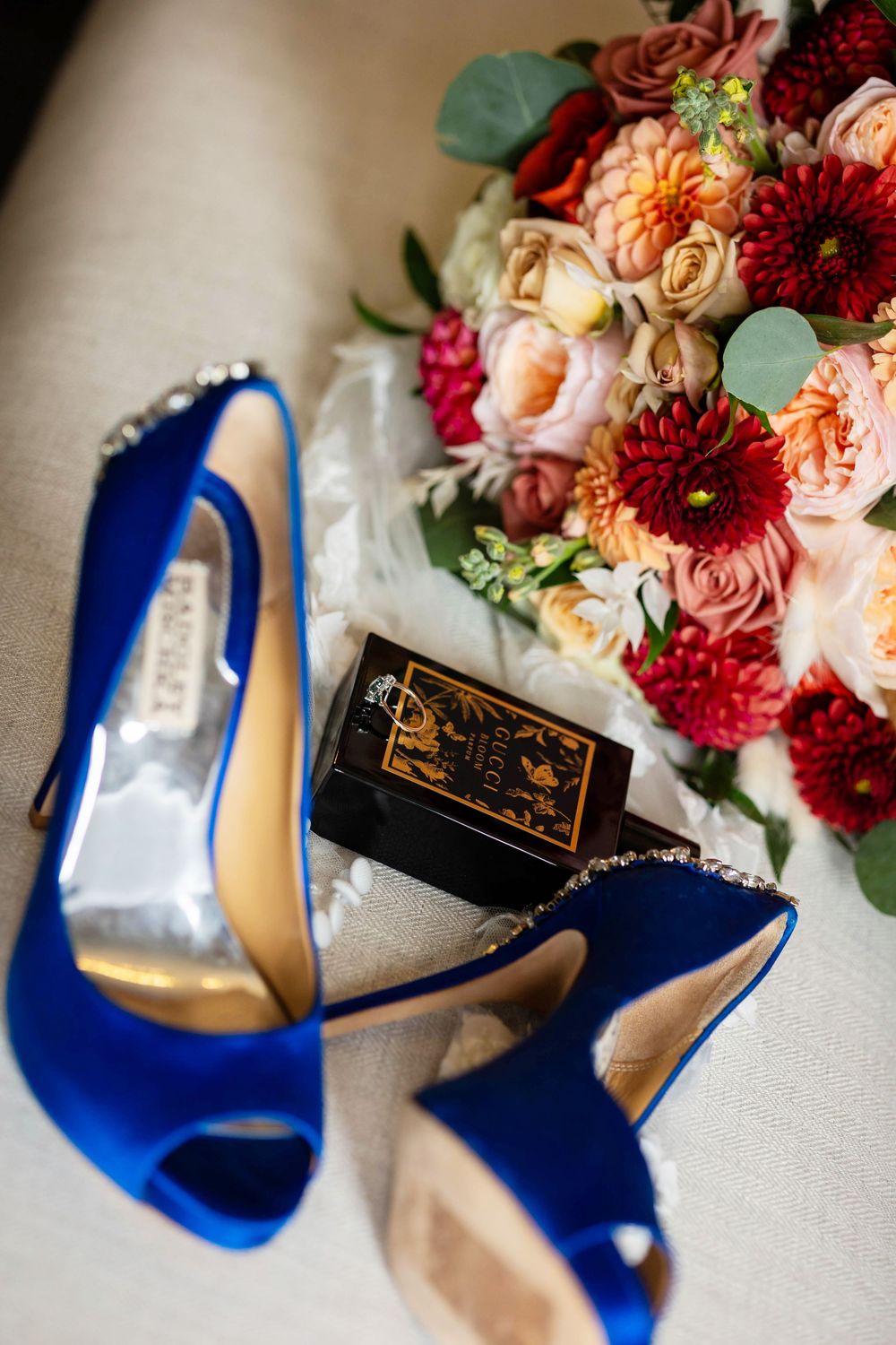 Elegant royal blue high heel shoes arranged with vibrant red and peach floral bouquet on light beige surface.